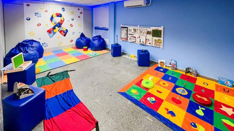 Imagem de uma sala com fundo azul, com o logotipo do autismo, com brinquedos e espaços sensoriais coloridos